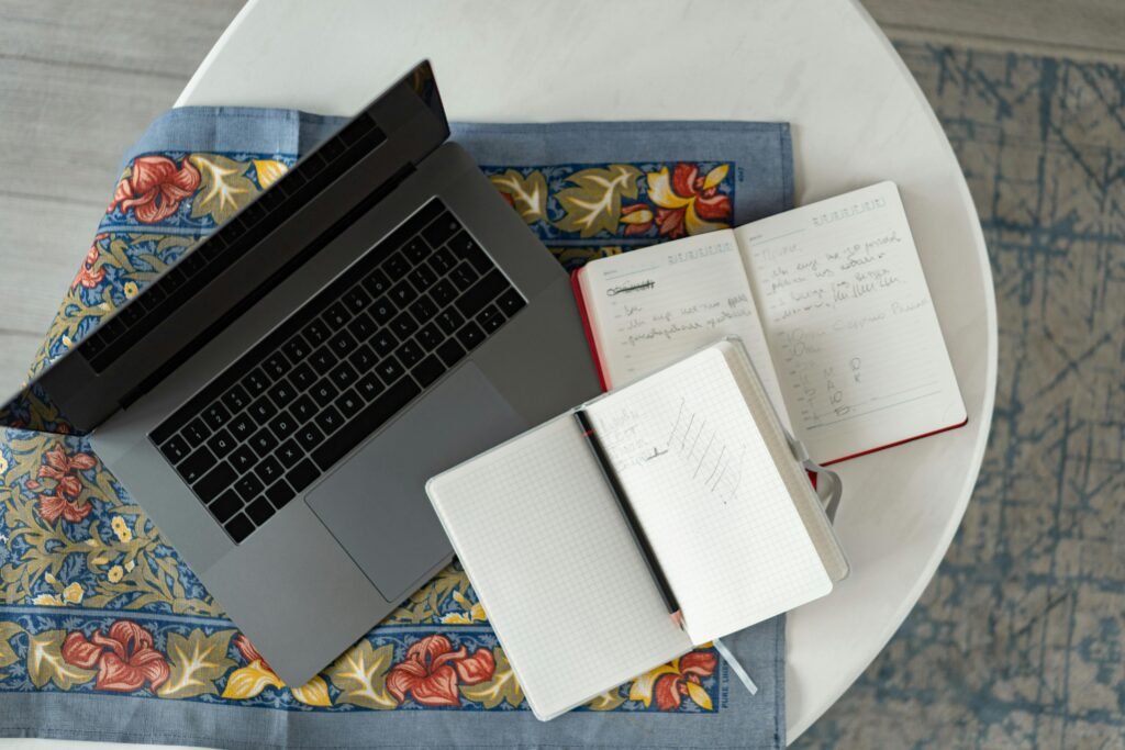 Overhead shot of a laptop with notebooks and a pencil on a patterned cloth.